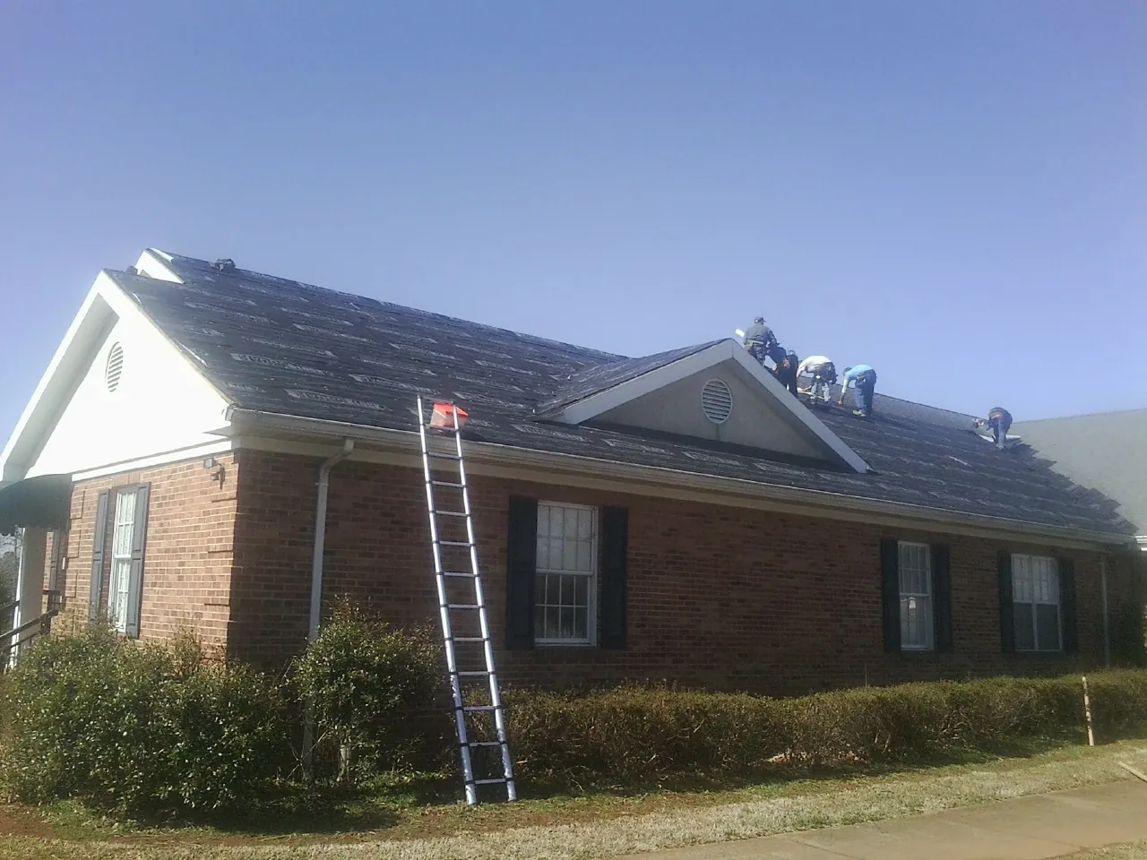 Crew tearing shingles off a brick ranch roof with a ladder set — Metal Roof Repair work by our crew in Currituck