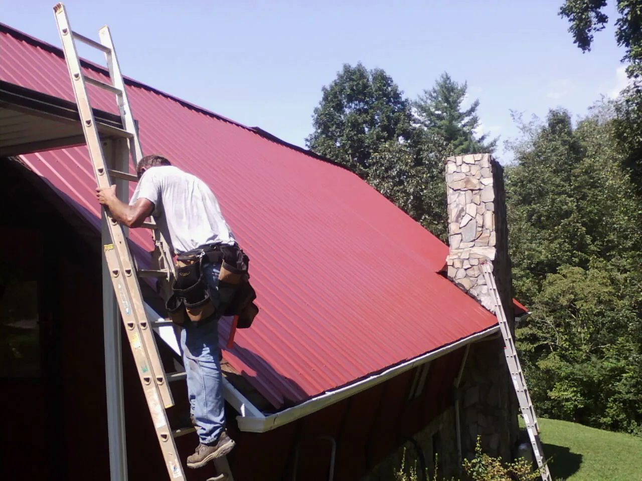 Roofer on a ladder beside a red metal roof and stone chimney — Soffit & Fascia Repair by Roofing Brothers in Currituck